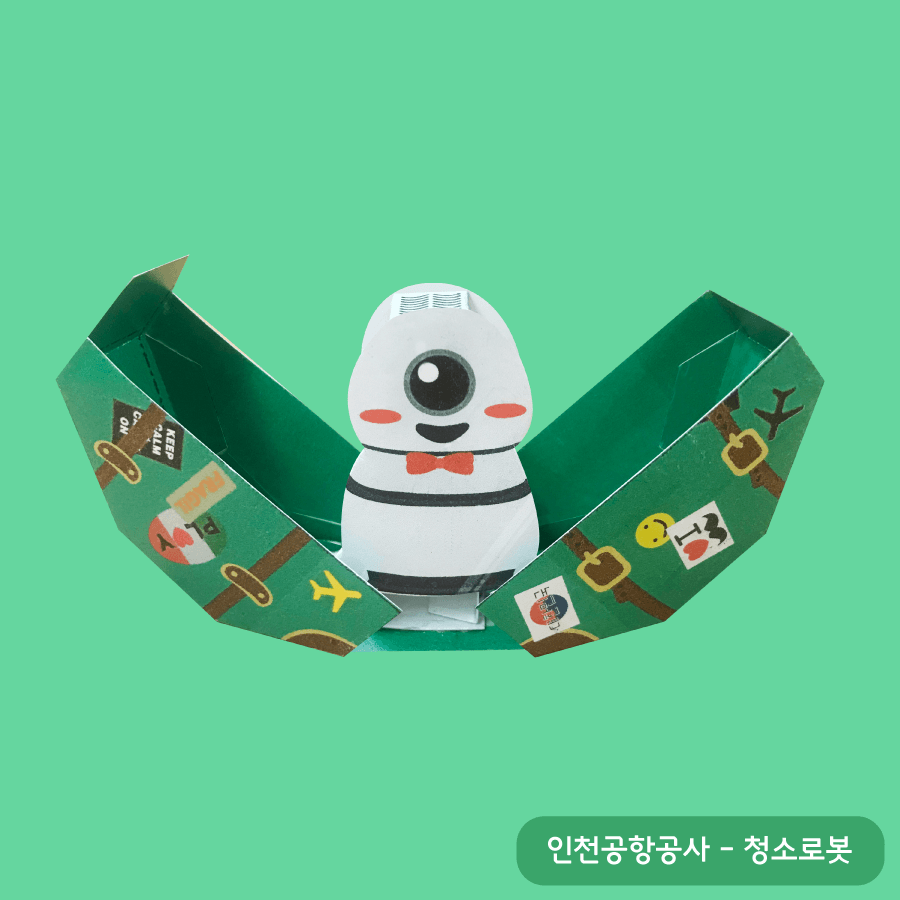 여행 스티커가 붙은 초록 캐리어가 양쪽으로 활짝 열리며 빨간 나비넥타이를 맨 외눈 청소로봇이 나타난다
