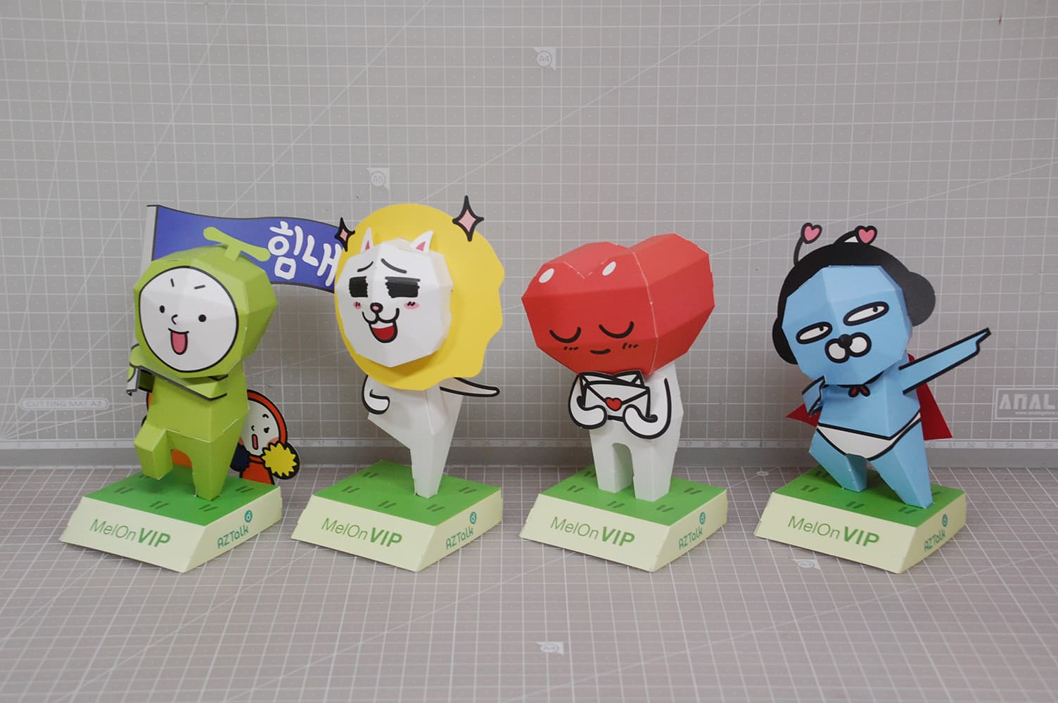 깃발을 든 초록 캐릭터와 노란 원반 뒤 흰 고양이, 하트 머리, 망토 두른 파란 캐릭터가 MelOn VIP 받침대에 선 페이퍼토이