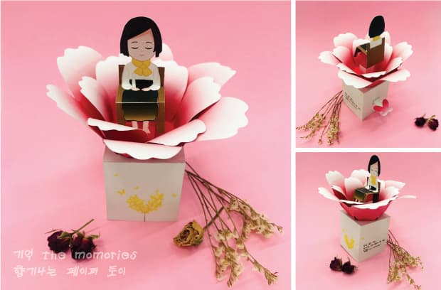 분홍 배경에 마른 꽃다발과 함께 놓인 소녀상 페이퍼토이, 활짝 벌어진 꽃잎 안에 의자에 앉은 단발머리 소녀가 있다