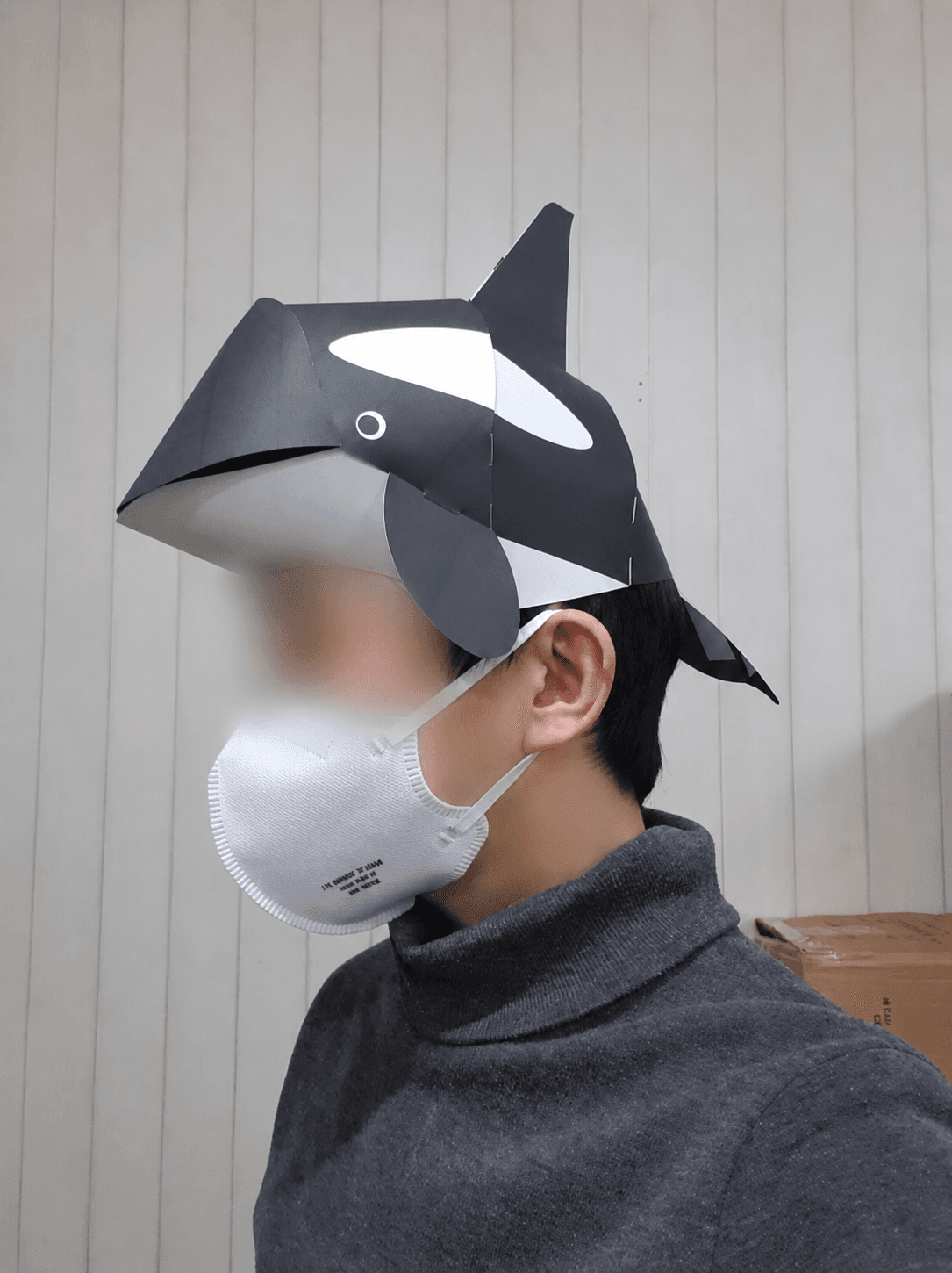 회색 니트를 입은 사람이 범고래 종이 모자를 쓰고 옆을 향해 서 있고 꼬리지느러미가 뒤통수 뒤로 뻗어 있어요
