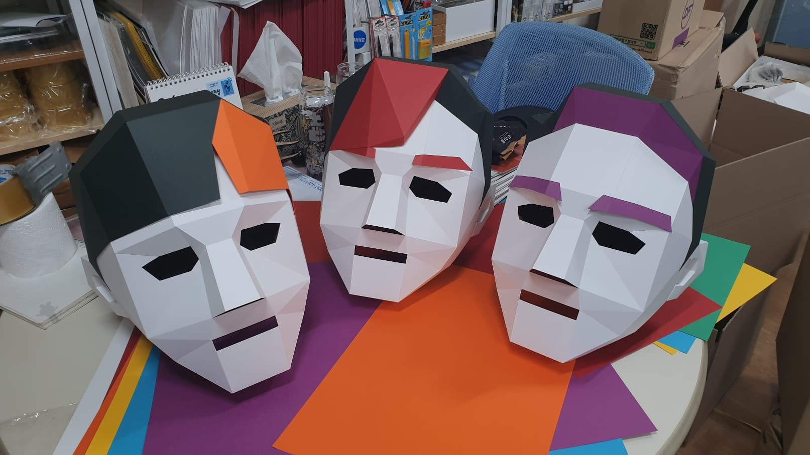검정·빨강·보라 머리를 얹은 로우폴리 흰 얼굴 종이 가면 세 개, 눈과 입이 뚫려 있어 쓰고도 앞을 볼 수 있다