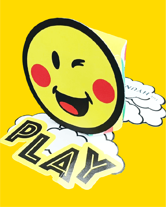 노란 배경 위로 윙크하며 웃는 스마일리 얼굴이 솟아 있고, 구름 모양 받침에 PLAY 레터링이 붙은 입체 종이 굿즈