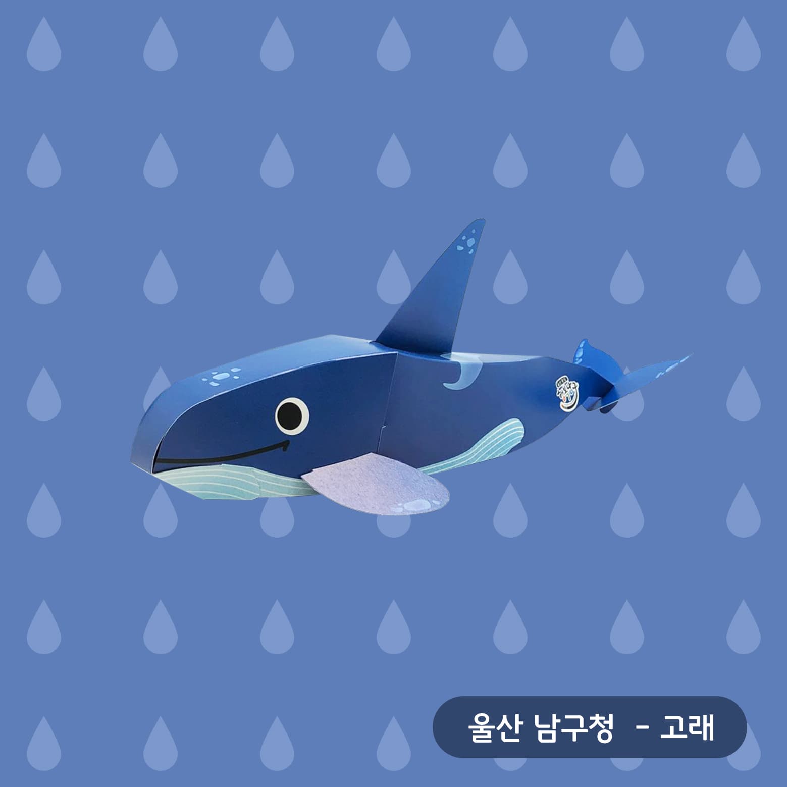 물방울 무늬 남색 배경 위, 웃는 입과 큰 눈에 등지느러미를 세운 파란 고래 페이퍼토이가 헤엄치듯 놓여 있다