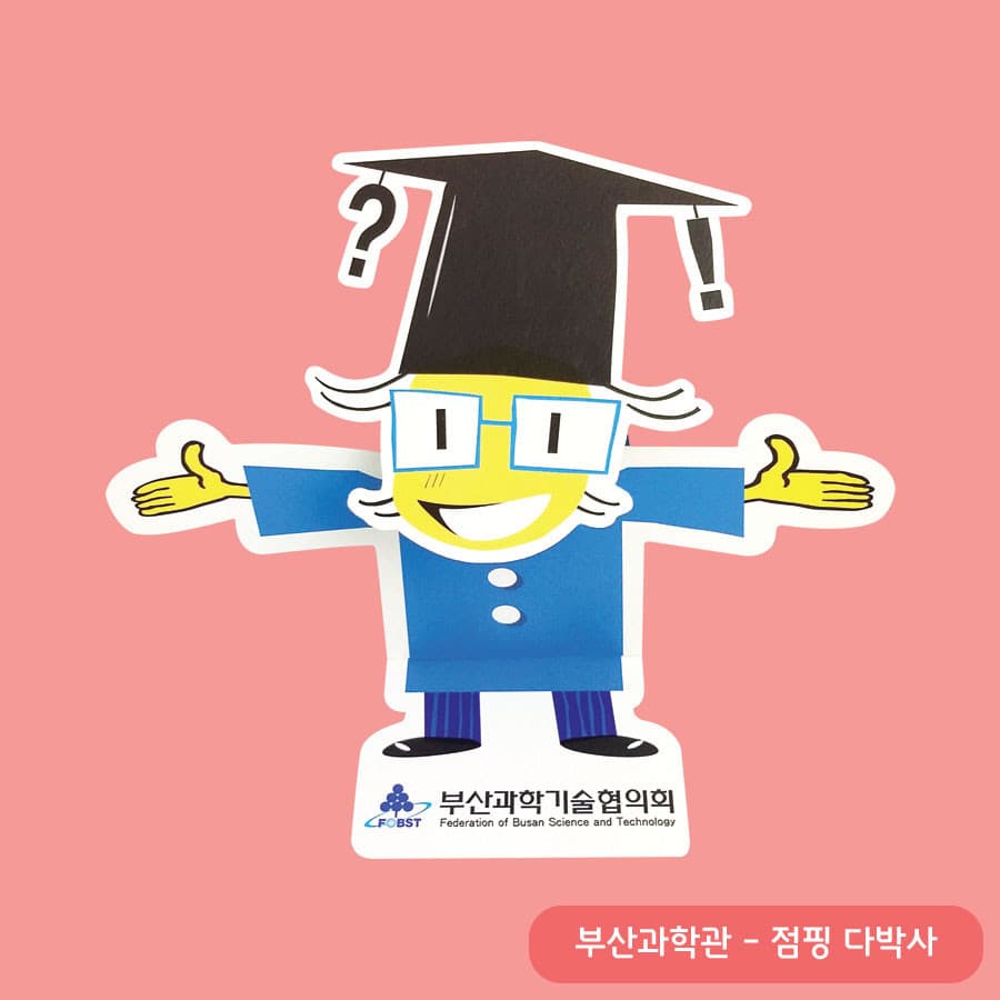 물음표와 느낌표가 달린 학사모를 쓰고 파란 안경 너머로 활짝 웃으며 두 팔을 벌린 다박사 페이퍼 토이, 받침에 부산과학기술협의회 로고
